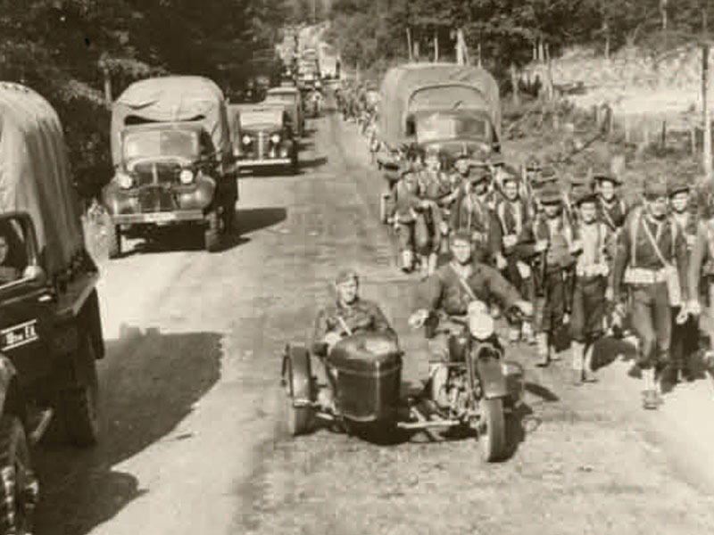 Black and white photograph of a group of men walking alongside a road lined with vehicles.